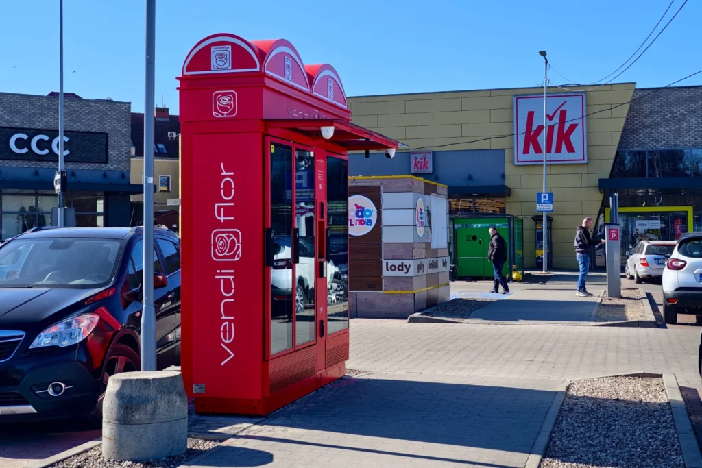 Red VendiFlor flower vending machine located in a retail park in Bolesławiec, visible from pedestrian walkway and parking area